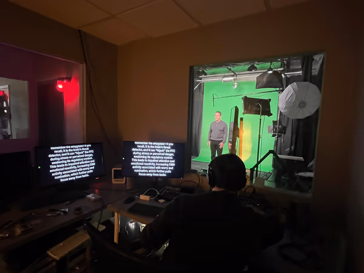 Person operating a video editing station with dual monitors displaying text, watching a man standing in front of a green screen in a studio with lighting equipment.