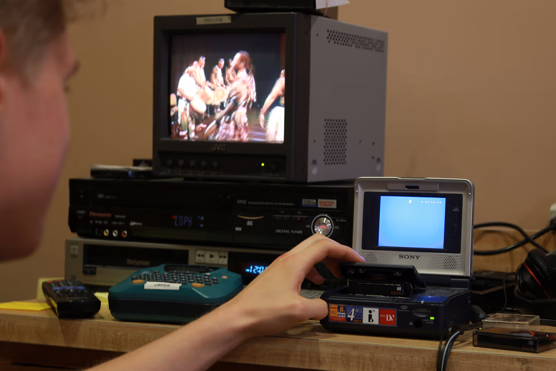 Person inserting a tape into a Sony Mini DV camcorder with a preview monitor and media equipment on a wooden table.