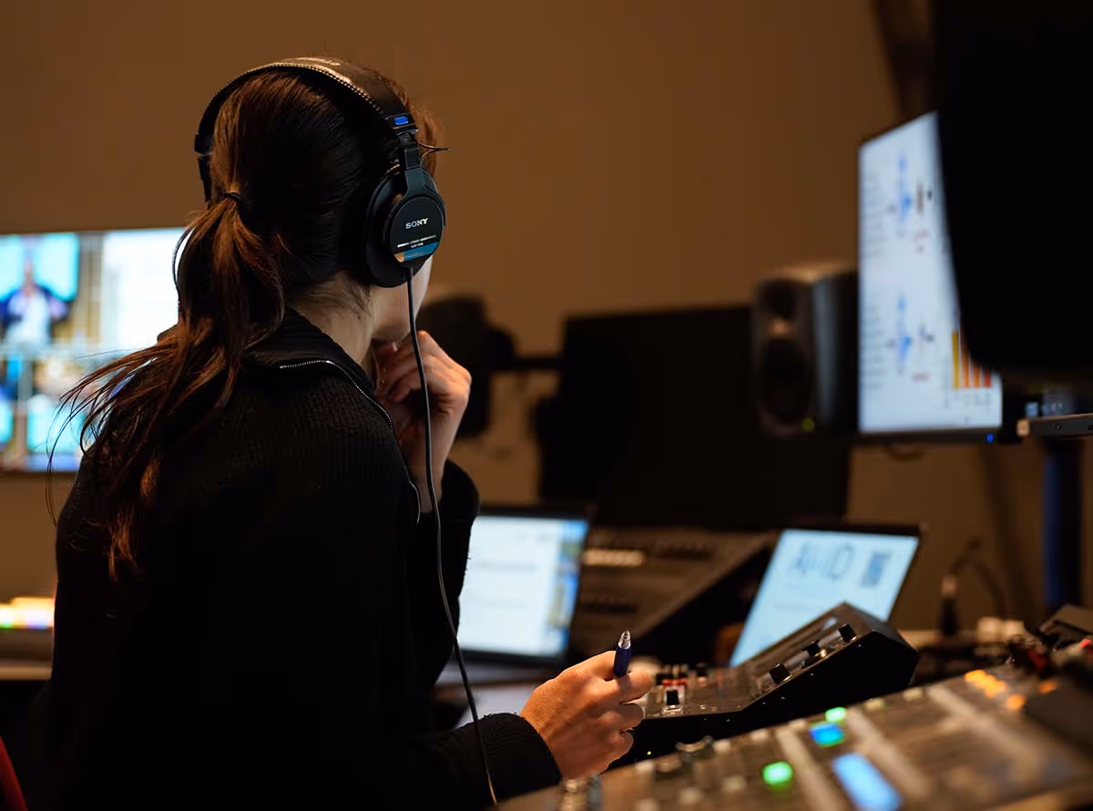 Person wearing headphones and working at an audio mixing console with multiple screens and controls in a studio.