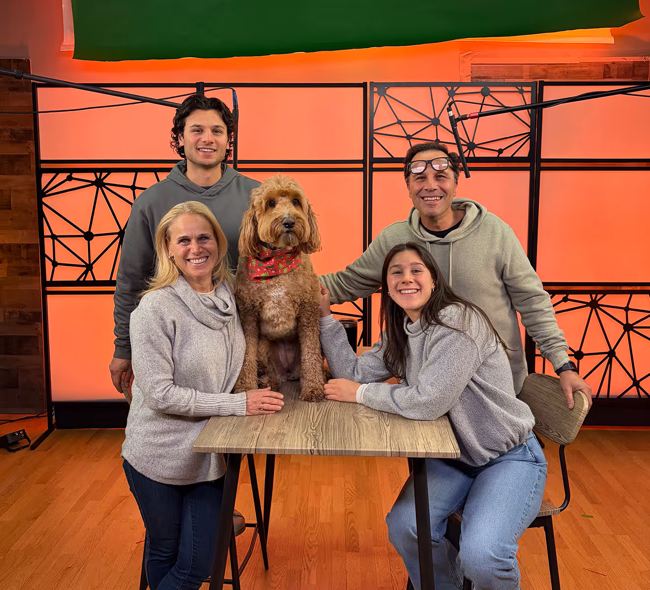 Group of four people smiling around a dog sitting on a wooden table against an orange and black geometric background.