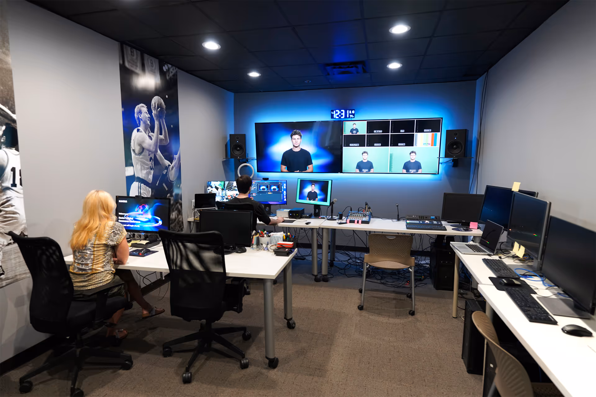Control room with multiple computer monitors, two people working, and a large screen wall displaying video feeds.