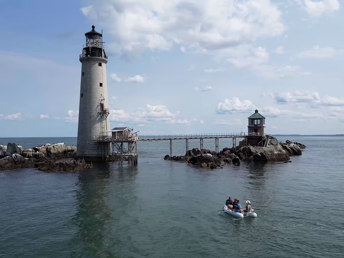 Small boat with four people rowing near a tall lighthouse connected by a narrow pier to a smaller structure on rocky shore.