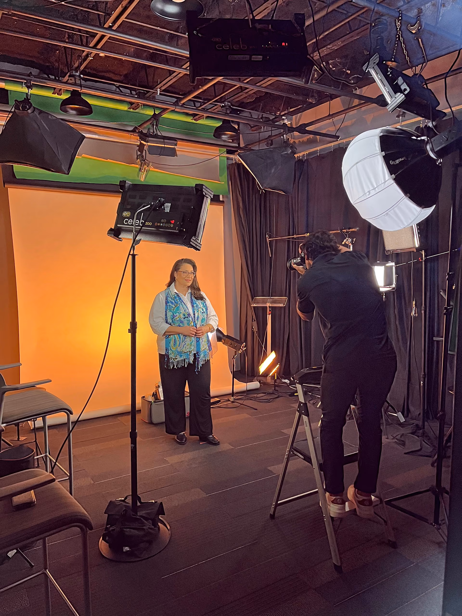 A woman with long dark hair wearing glasses, a blue scarf, and white shirt stands in a photo studio with an orange backdrop, surrounded by professional lighting equipment and a photographer who is taking her picture while standing on a ladder.