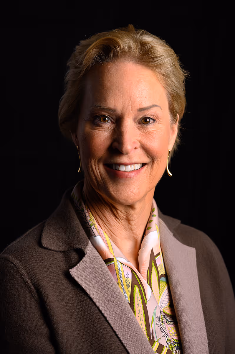 Smiling middle-aged woman with short blonde hair wearing gold earrings, floral scarf, and dark brown jacket against a black background.