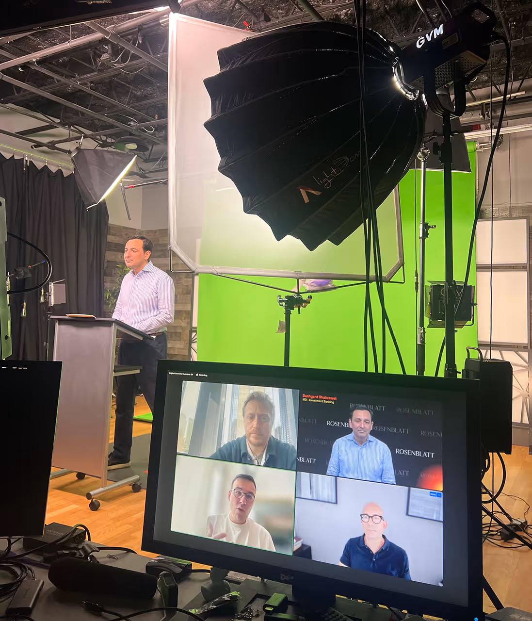 Man standing at a podium in a studio with green screen and lighting equipment, while a monitor in the foreground shows a video call with four participants.