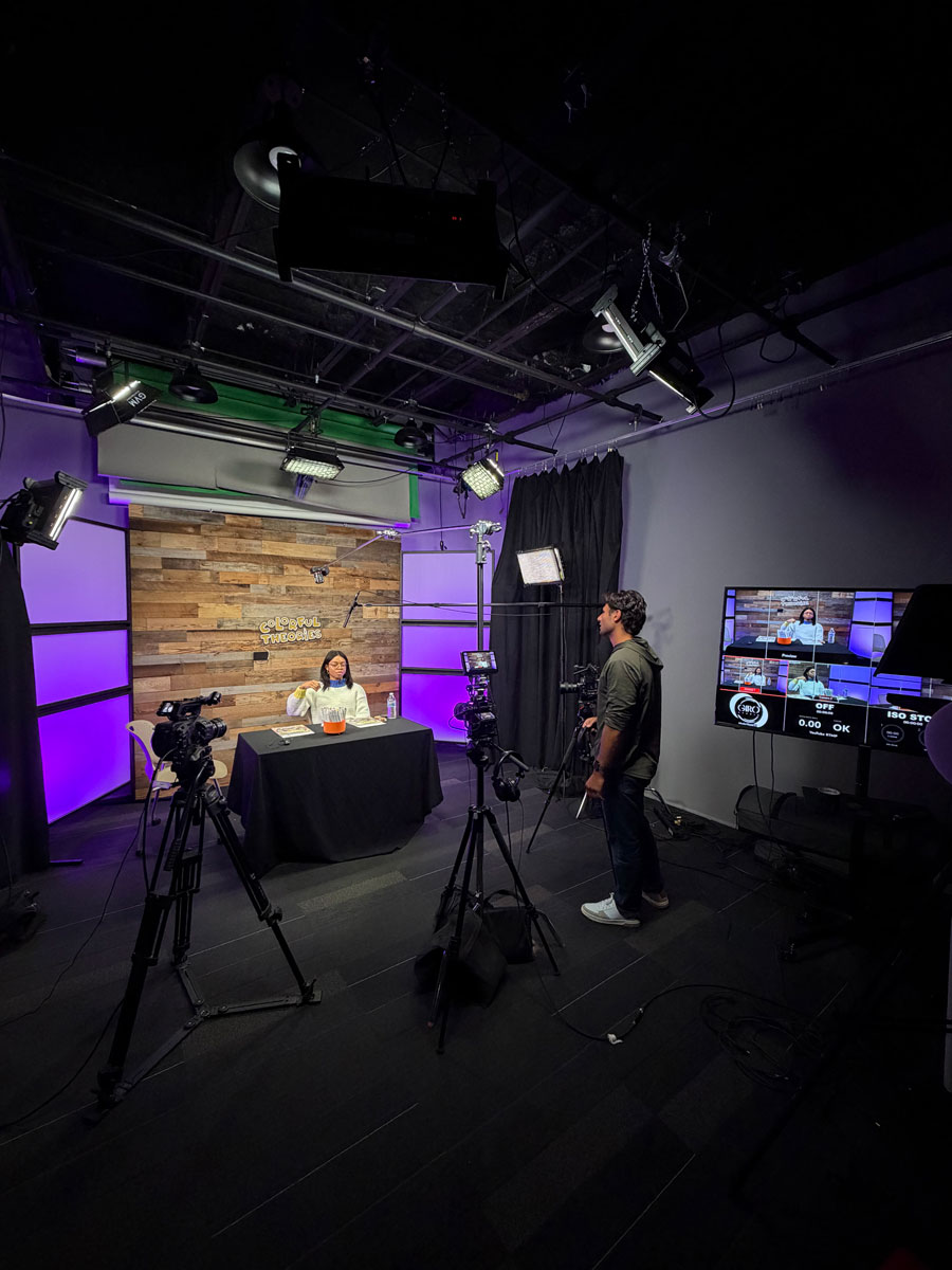 A video production studio with a woman seated at a table labeled 'Colorful Theories' on a wooden backdrop, surrounded by cameras, lights, and a man operating a camera while a monitor shows multiple camera angles.