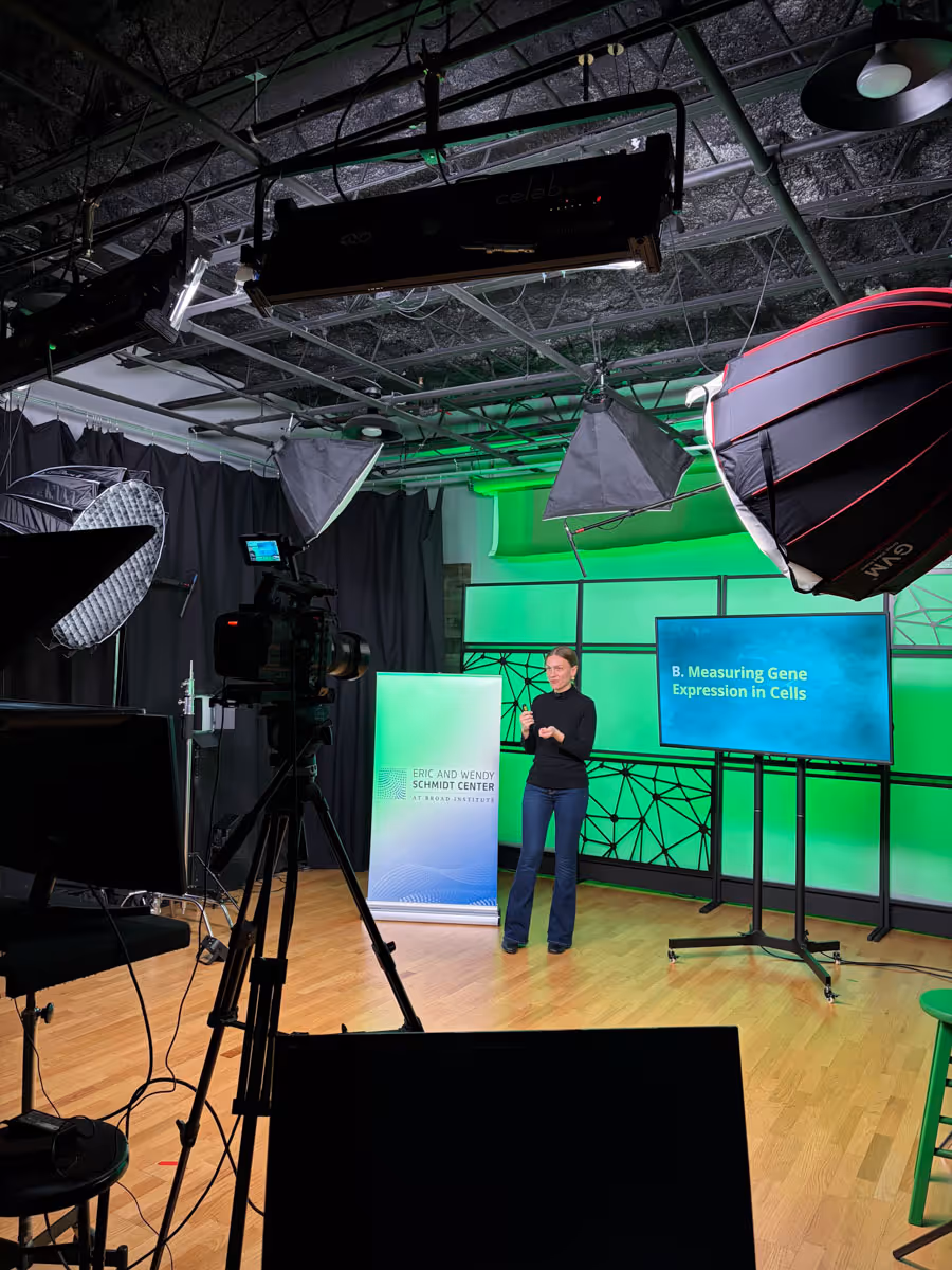 A woman standing in a studio with professional lighting and cameras, presenting in front of a green backdrop and a screen displaying 'B. Measuring Gene Expression in Cells.' A sign reads 'Eric and Wendy Schmidt Center.'