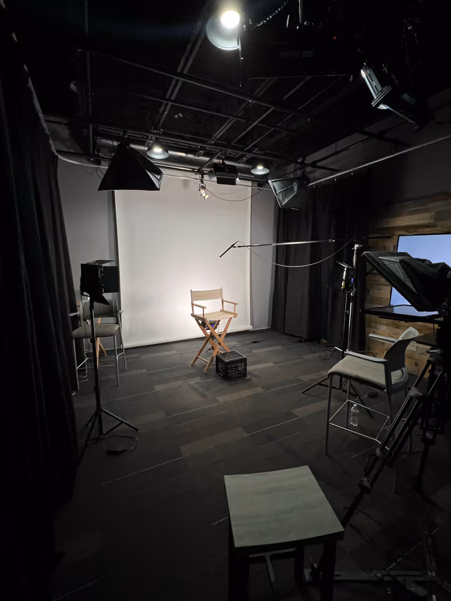 Indoor video or photo studio setup with a director's chair centered on a small black crate in front of a white backdrop, surrounded by lighting equipment, cameras, and chairs.