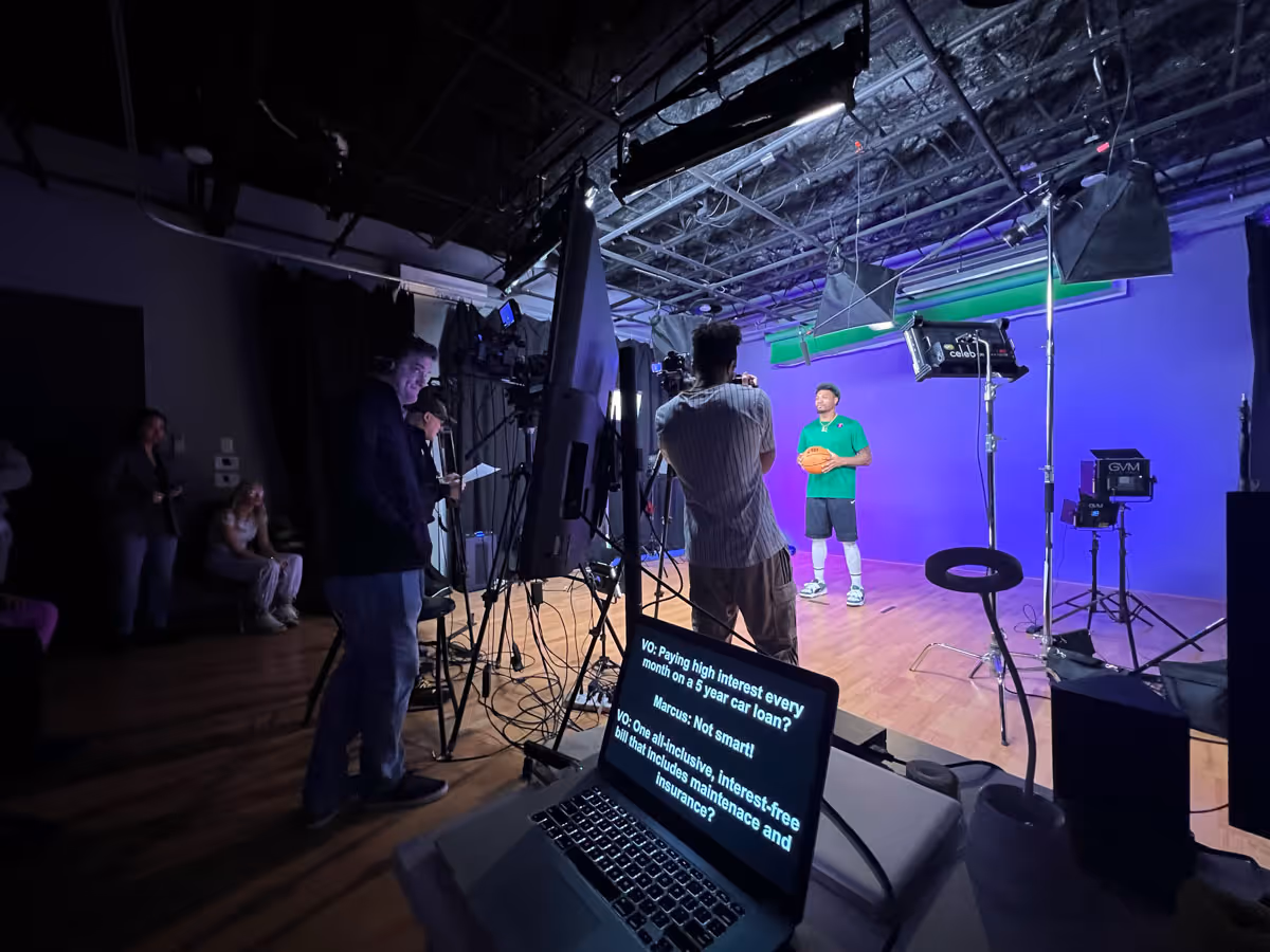 Behind-the-scenes view of a video production set with a man holding a basketball against a purple backdrop, surrounded by lighting and camera equipment, and crew members including a man facing the set and a laptop displaying script text.