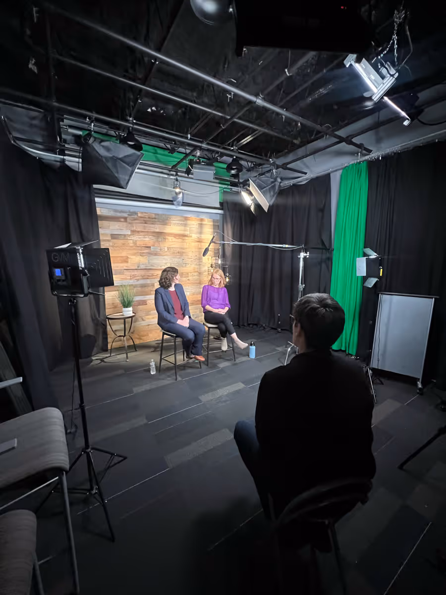 Two women seated on stools in a studio with wooden backdrop, engaged in conversation while a person in the foreground operates filming equipment, surrounded by professional lighting and boom microphones.