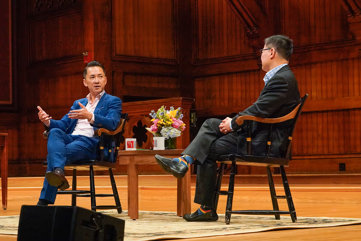 Two men seated on wooden chairs on a stage engaged in conversation with a small table with flowers and mugs between them.