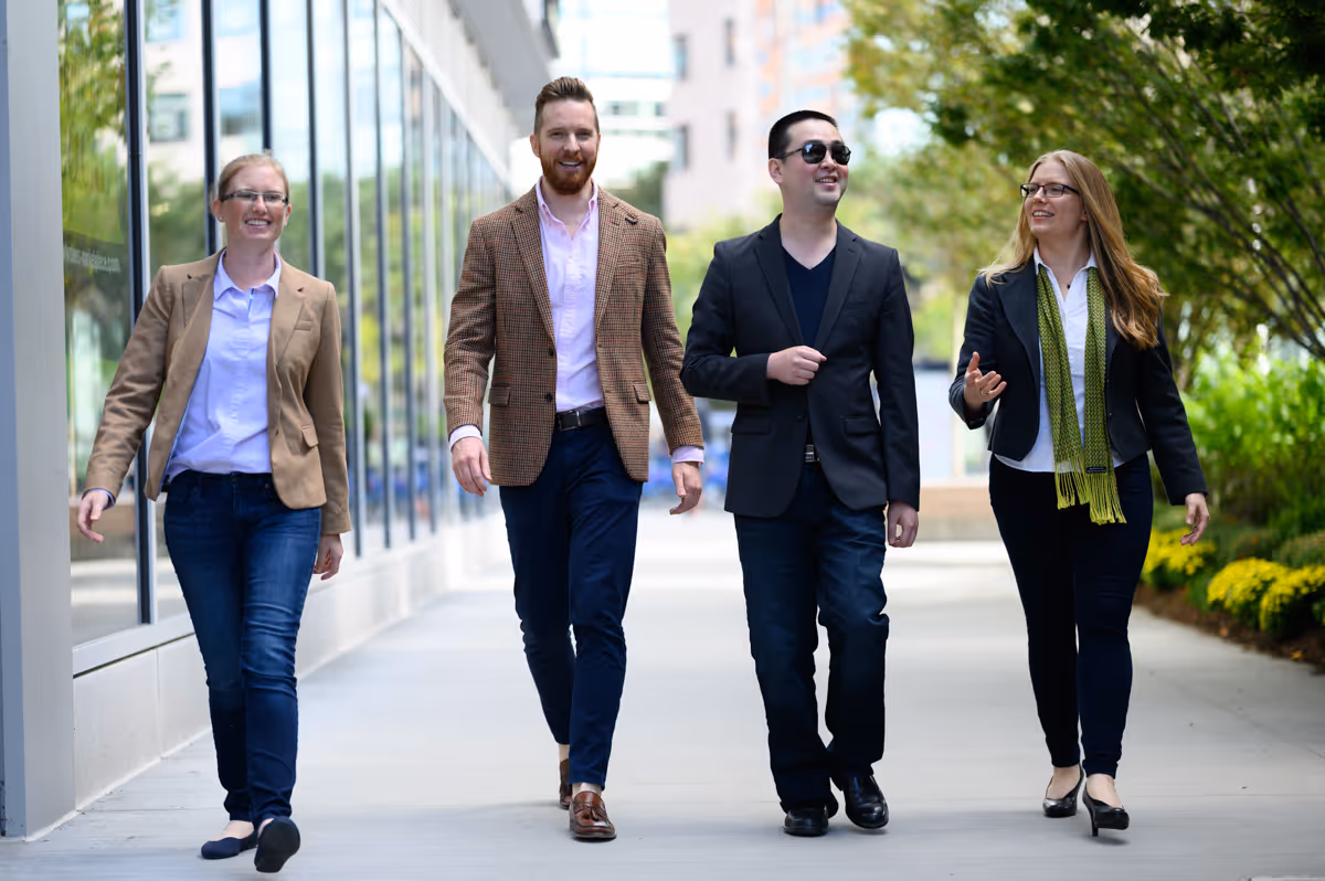 Four professionally dressed colleagues, two men and two women, walking outdoors on a city sidewalk and smiling.