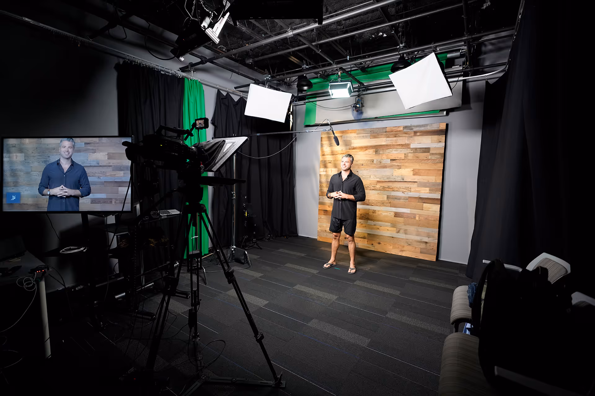 Man in black shirt and shorts standing in a professional video studio with camera, lighting, and a wooden backdrop.