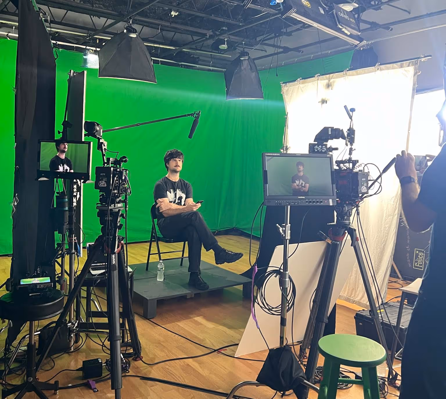 Man sitting on a chair in a green screen studio surrounded by professional video cameras, lighting equipment, and monitors.
