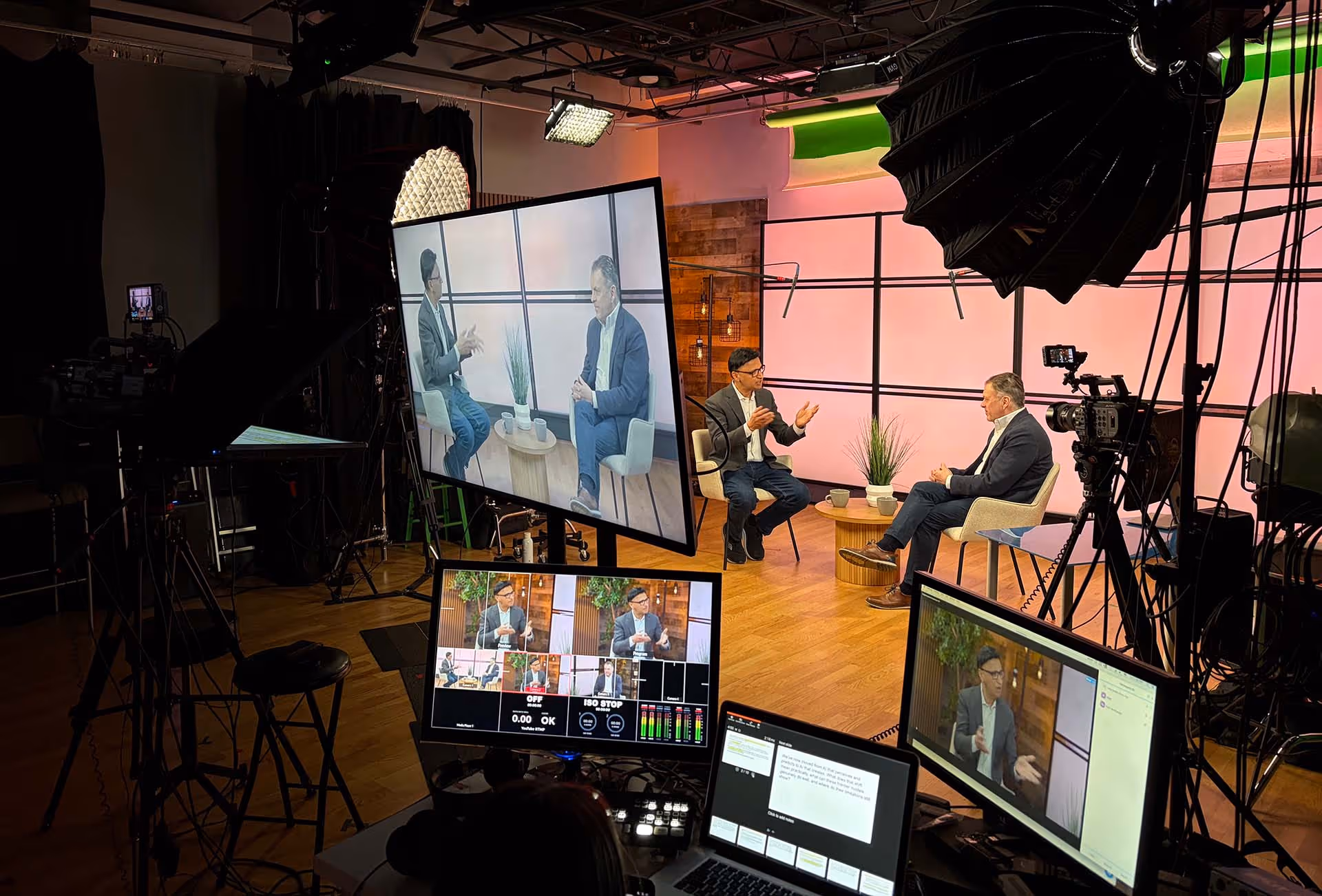 Studio setup showing two men seated and conversing on set with cameras, studio lights, monitors, and control screens in view.