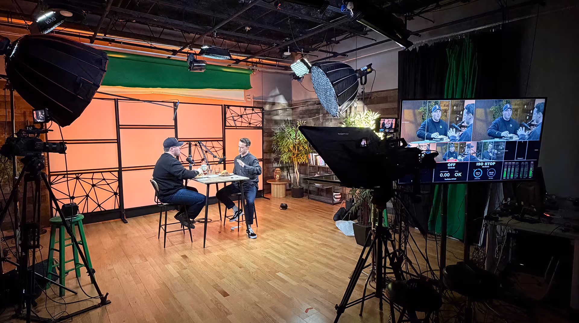 Two men seated at a high table in a professional studio with cameras, lighting equipment, and a monitor showing their live video feed.