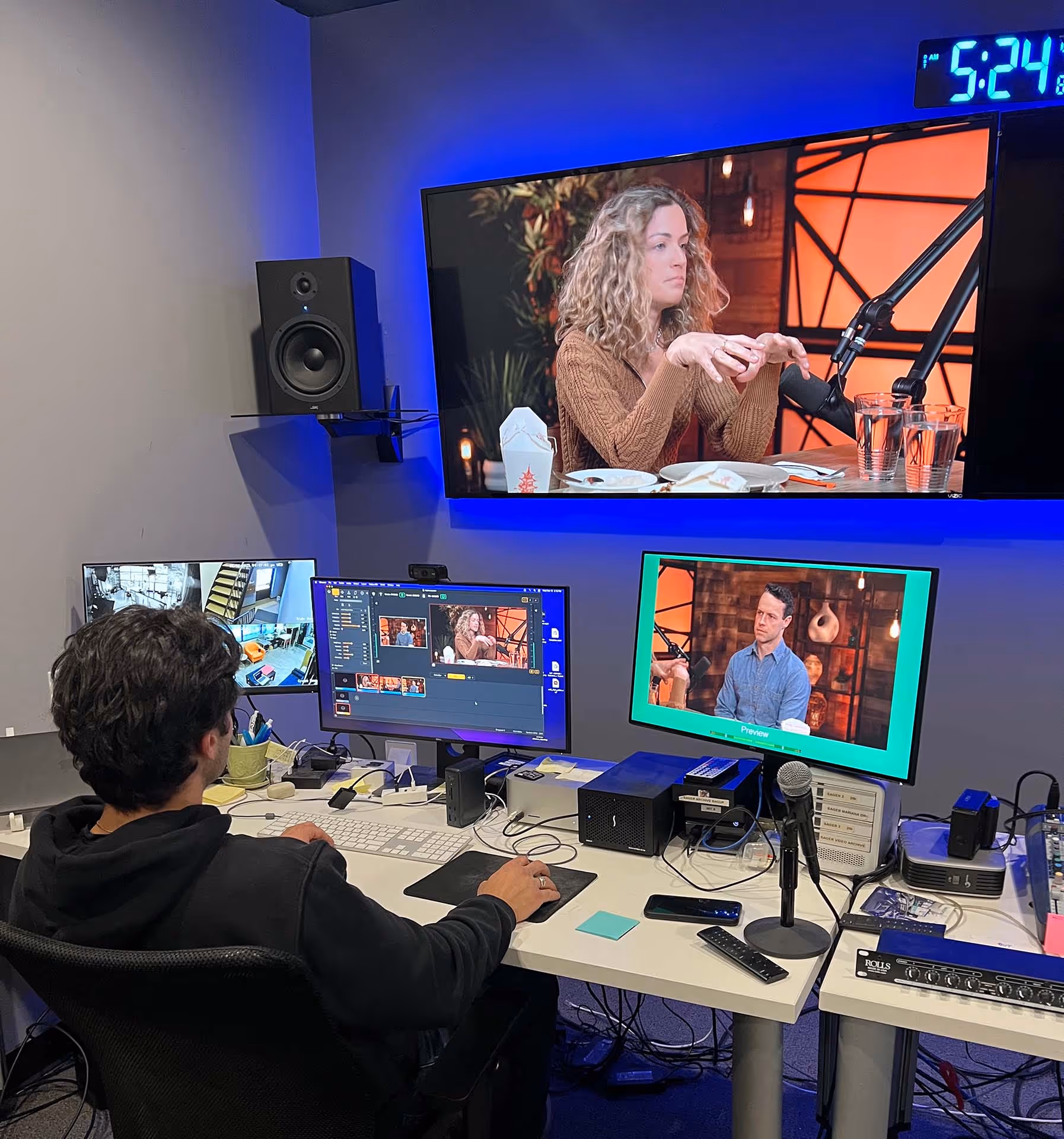Media control room with a person editing a video podcast on multiple monitors, showing a man and woman speaking on the screens.