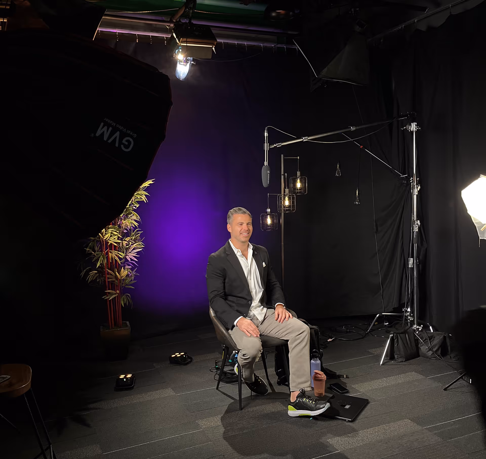 Man in a black blazer and light pants sitting on a chair in a studio with purple background lighting and professional filming equipment.