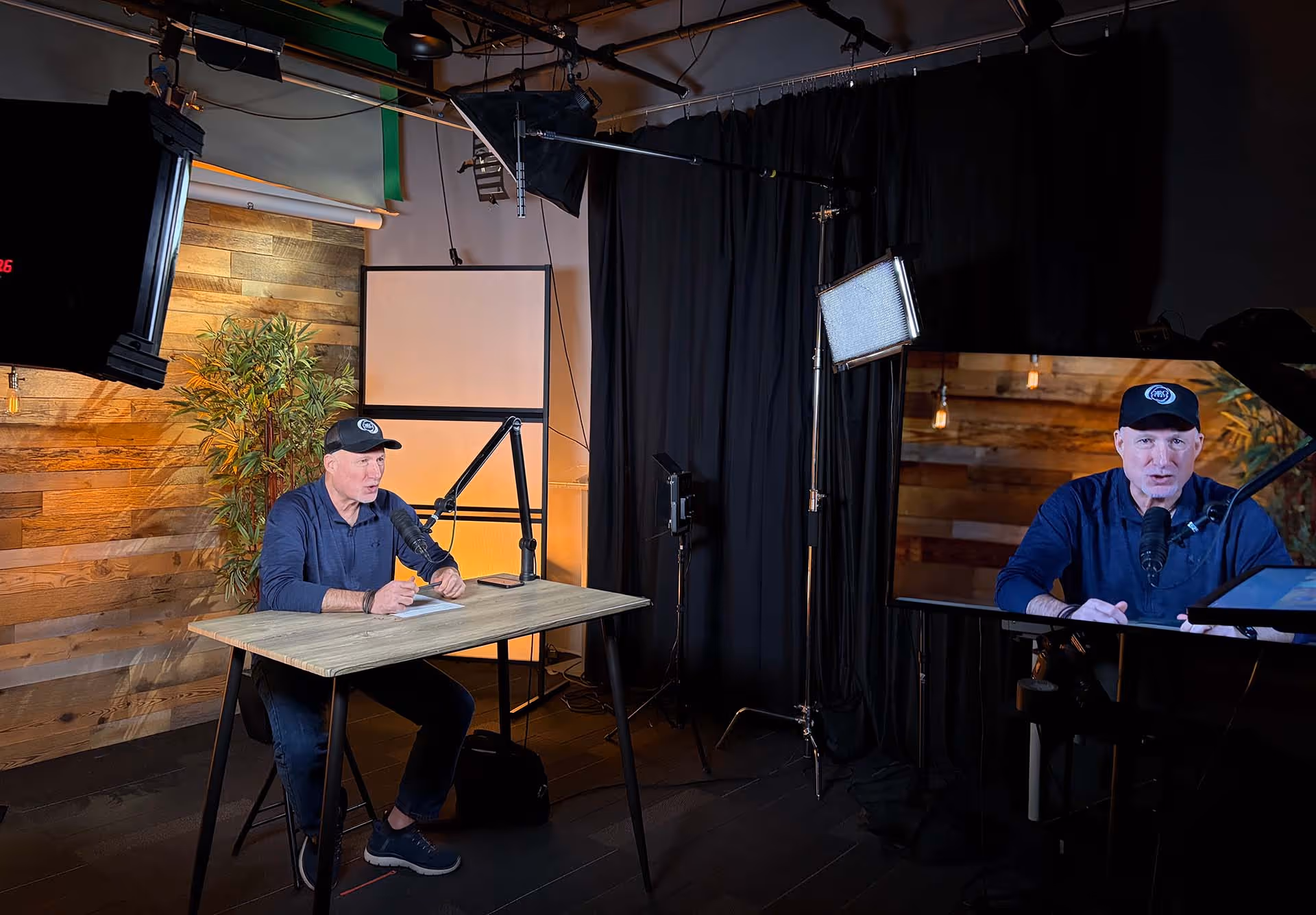 Man wearing a cap speaking into a microphone at a desk in a recording studio with professional lighting and a large monitor showing his live image.