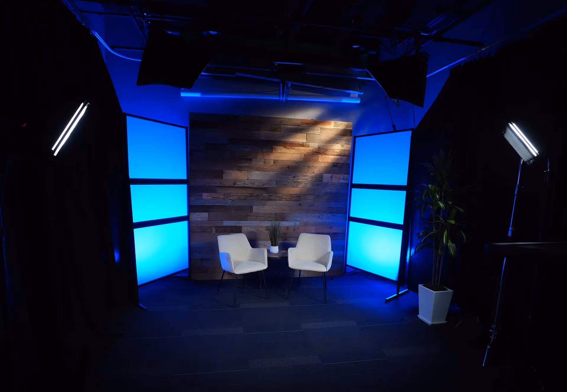 Studio setup with two white chairs, a small round table with a plant, wooden backdrop, blue illuminated panels, and studio lights.