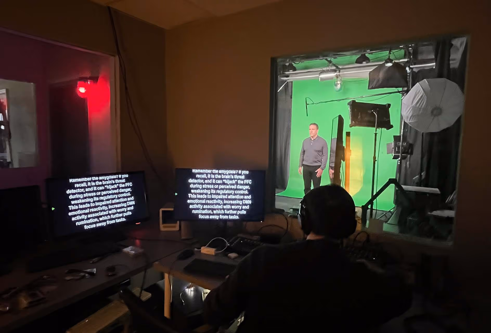 Man standing in front of a green screen in a video production studio, viewed from the control room with a person monitoring multiple screens.