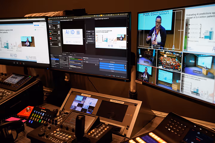 Control room with multiple monitors displaying video editing software, live video feeds, and a presenter discussing risk estimation.