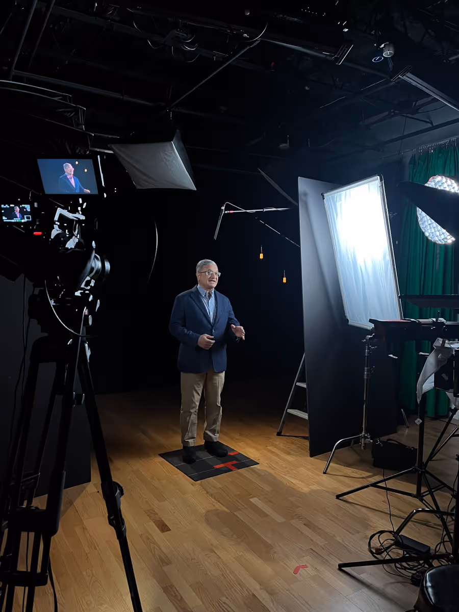 Man in a suit jacket and khaki pants standing on a marked floor square in a professional video studio, surrounded by lighting equipment, a boom microphone, and a camera on a tripod filming him.