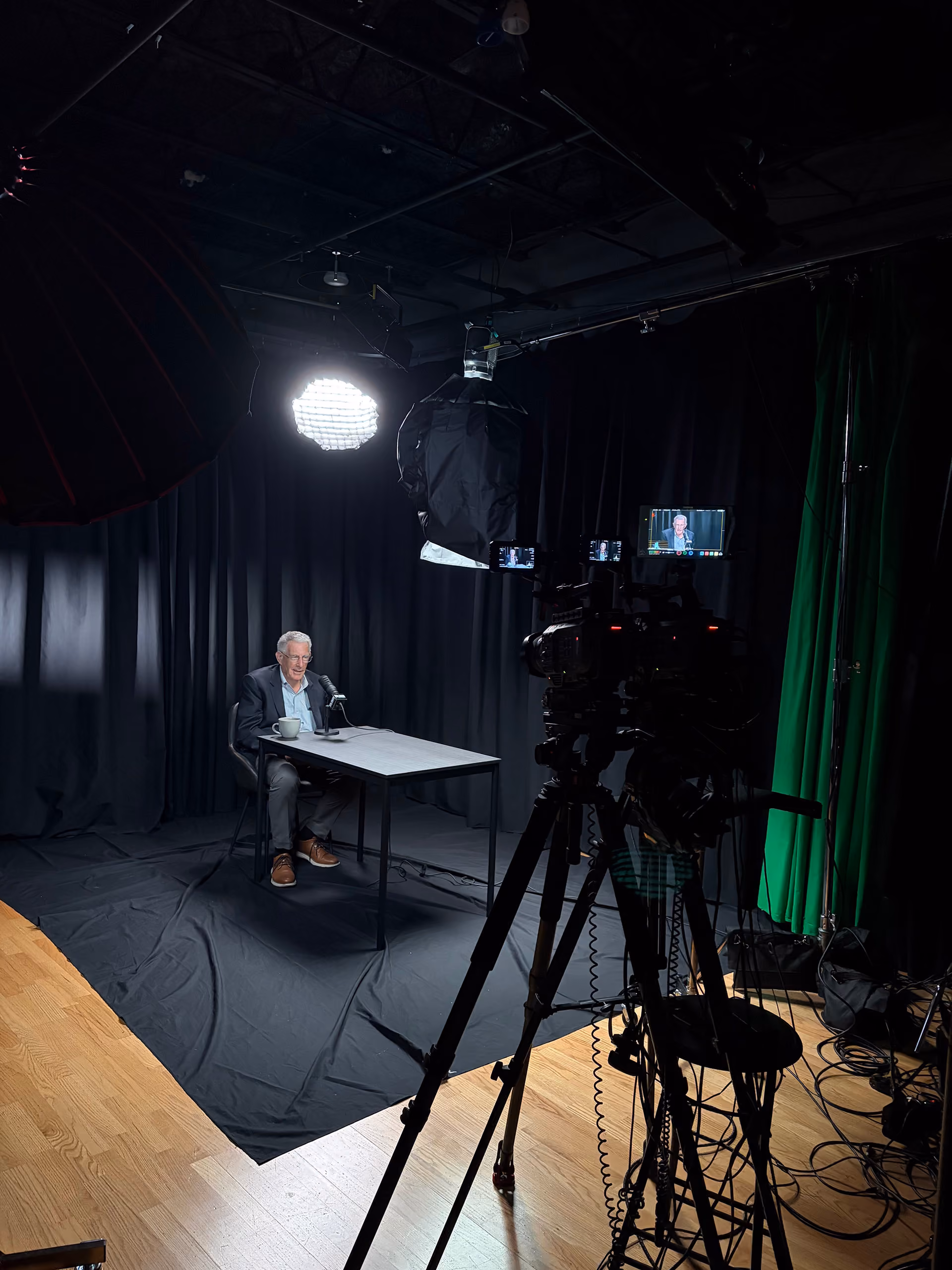 An older man in a suit sitting at a table with a microphone in a professional video recording studio with black curtains, studio lights, cameras on tripods, and green screen curtain visible.