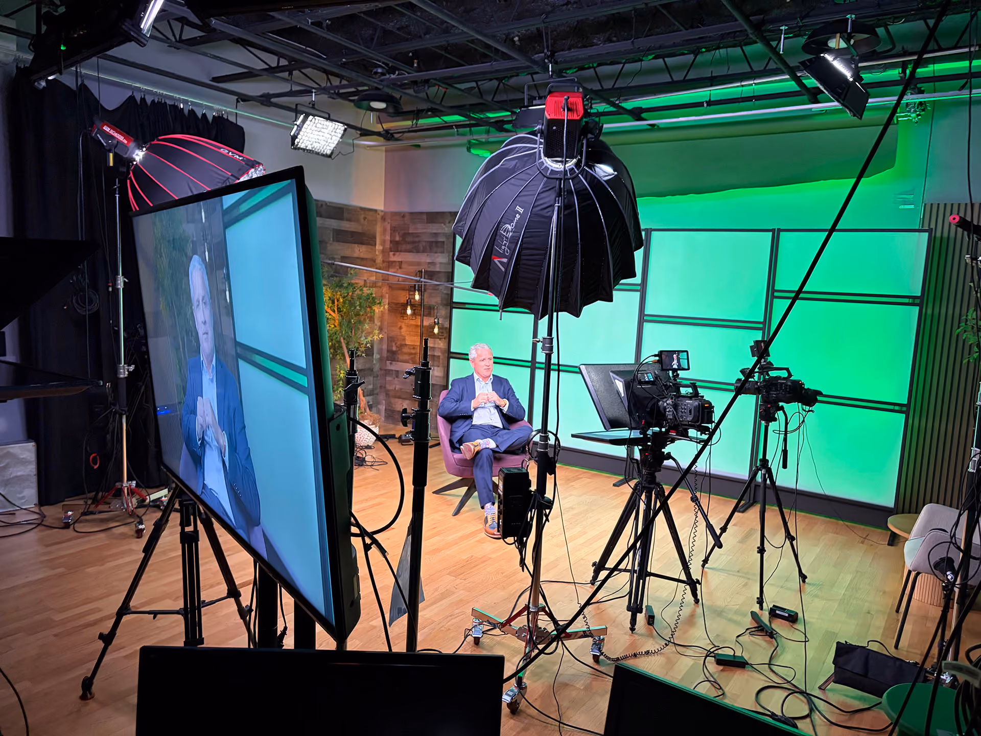 Professional video studio setup with a man seated on a chair in front of a green screen backdrop, surrounded by cameras, lighting equipment, and a large monitor showing his image.