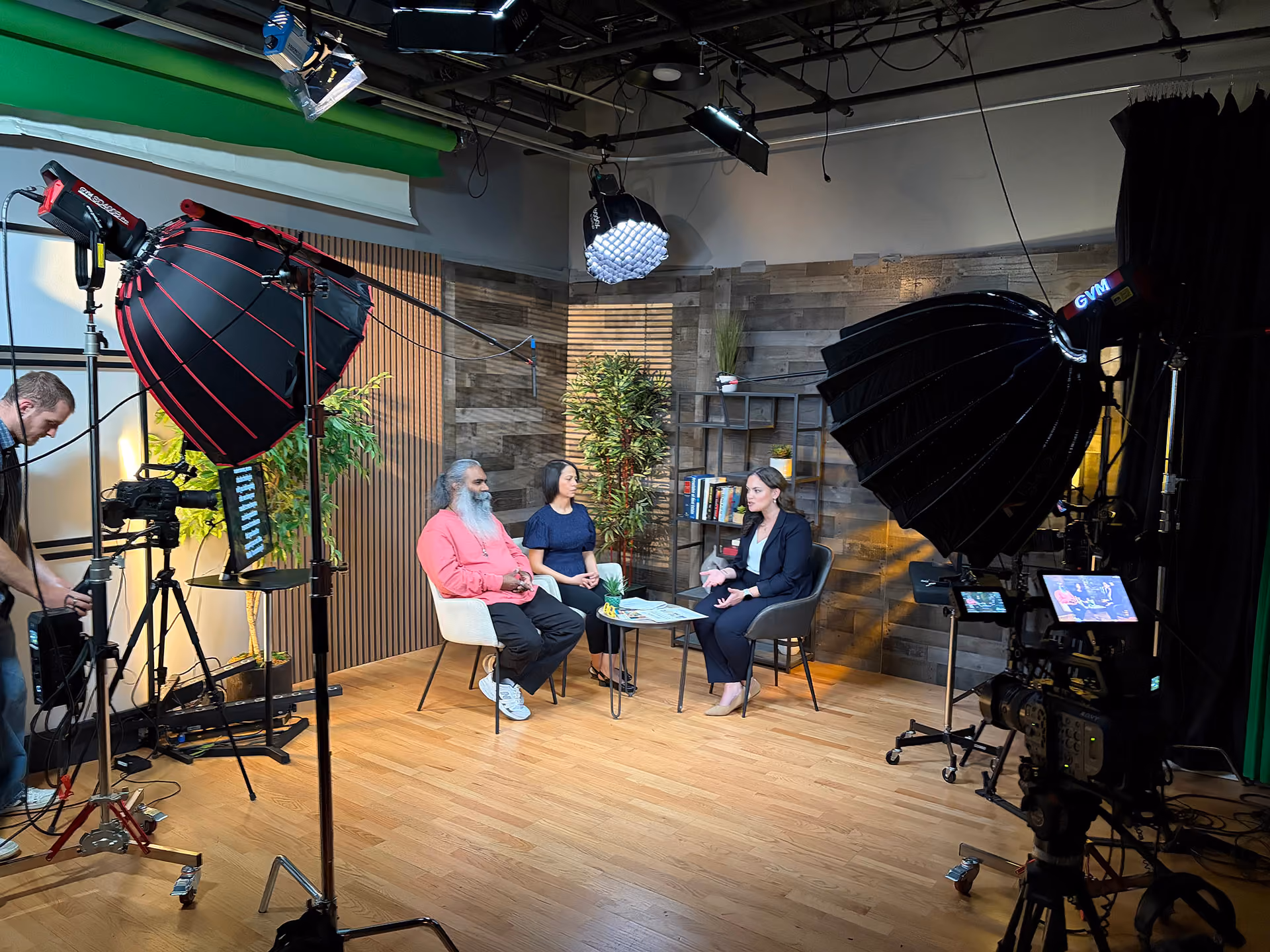 Three people seated in a studio with wood floors and a wood-paneled background, engaged in conversation around a small table, with professional lighting and camera equipment surrounding them and a crew member adjusting a camera on the left.
