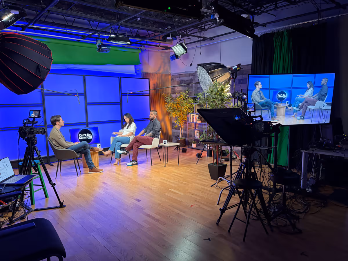 Three people seated on chairs having a discussion on a studio set with blue walls, soft lighting, cameras, and a large screen showing their live feed.