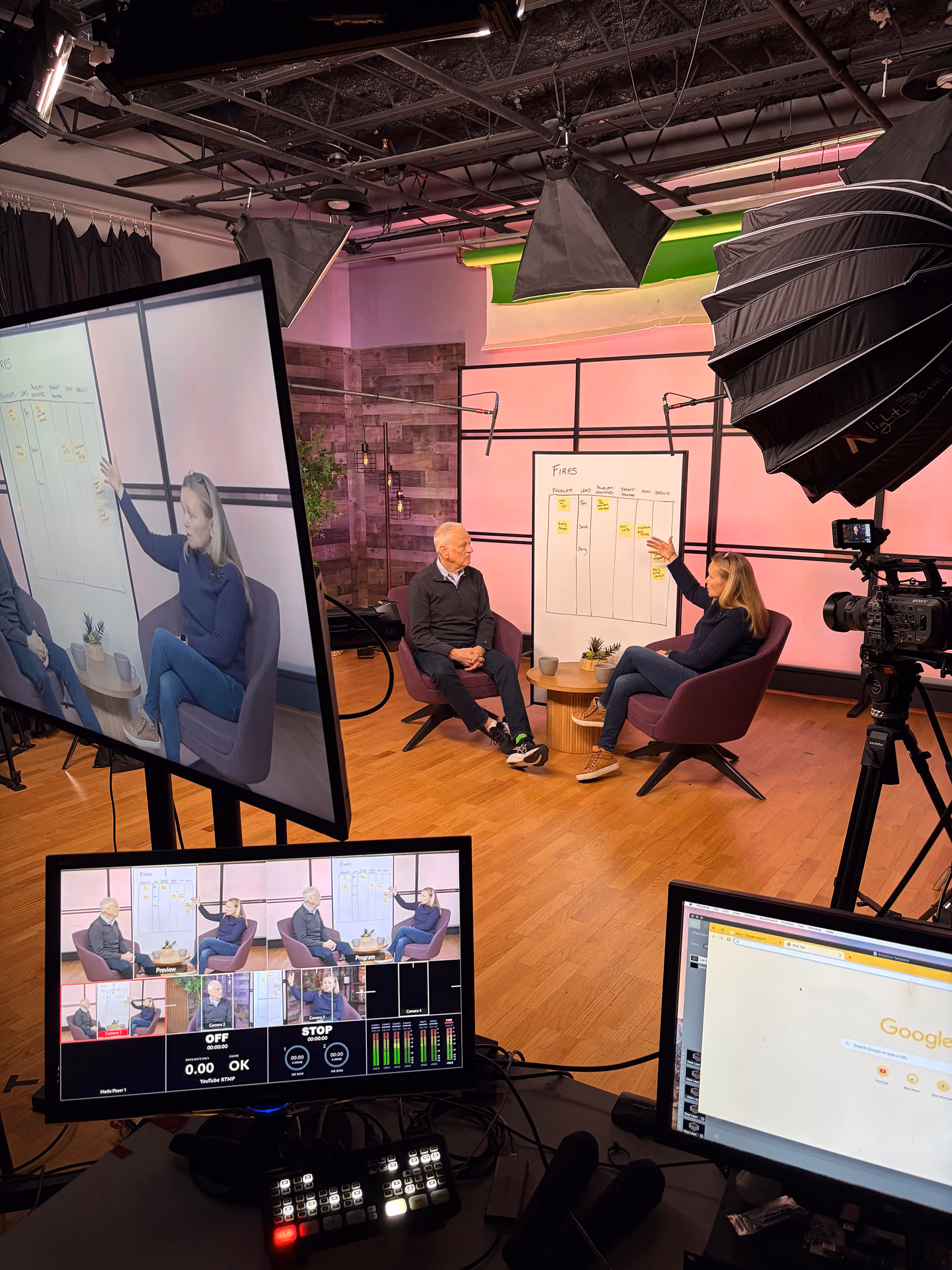 A video production studio with two people seated in purple chairs facing each other, next to a whiteboard with notes titled 'Fires'. Large studio lights and cameras are set up around them. Multiple monitors display the live video feed and controls of the recording.