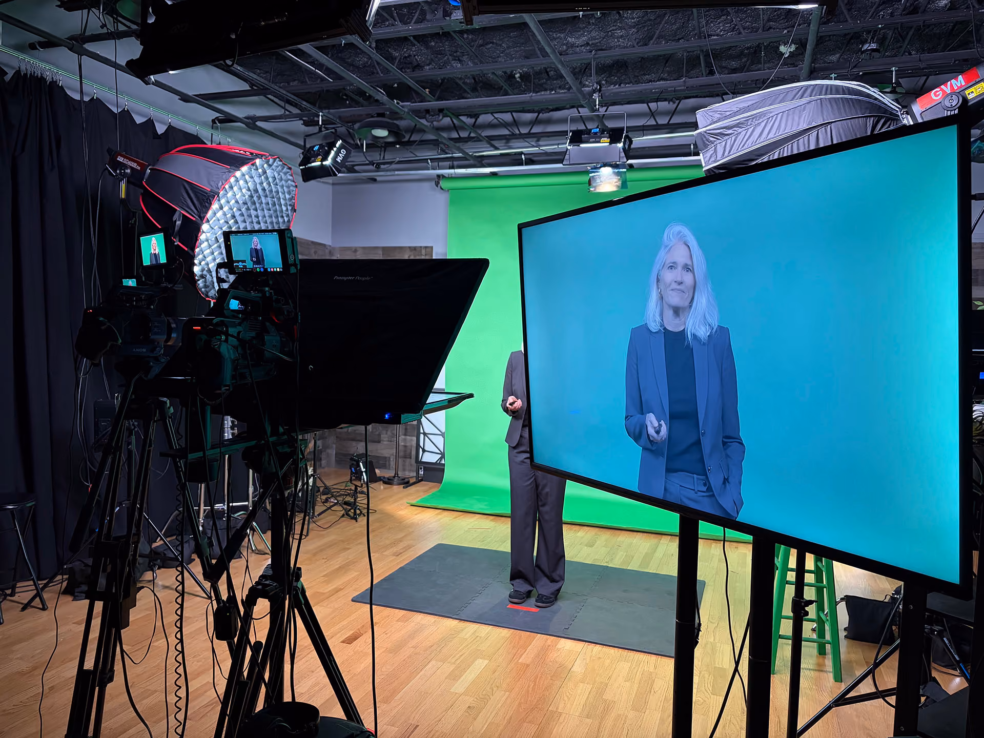 A woman with gray hair in a dark suit stands in front of a green screen in a video studio, with cameras, lighting equipment, and a large monitor displaying her image.