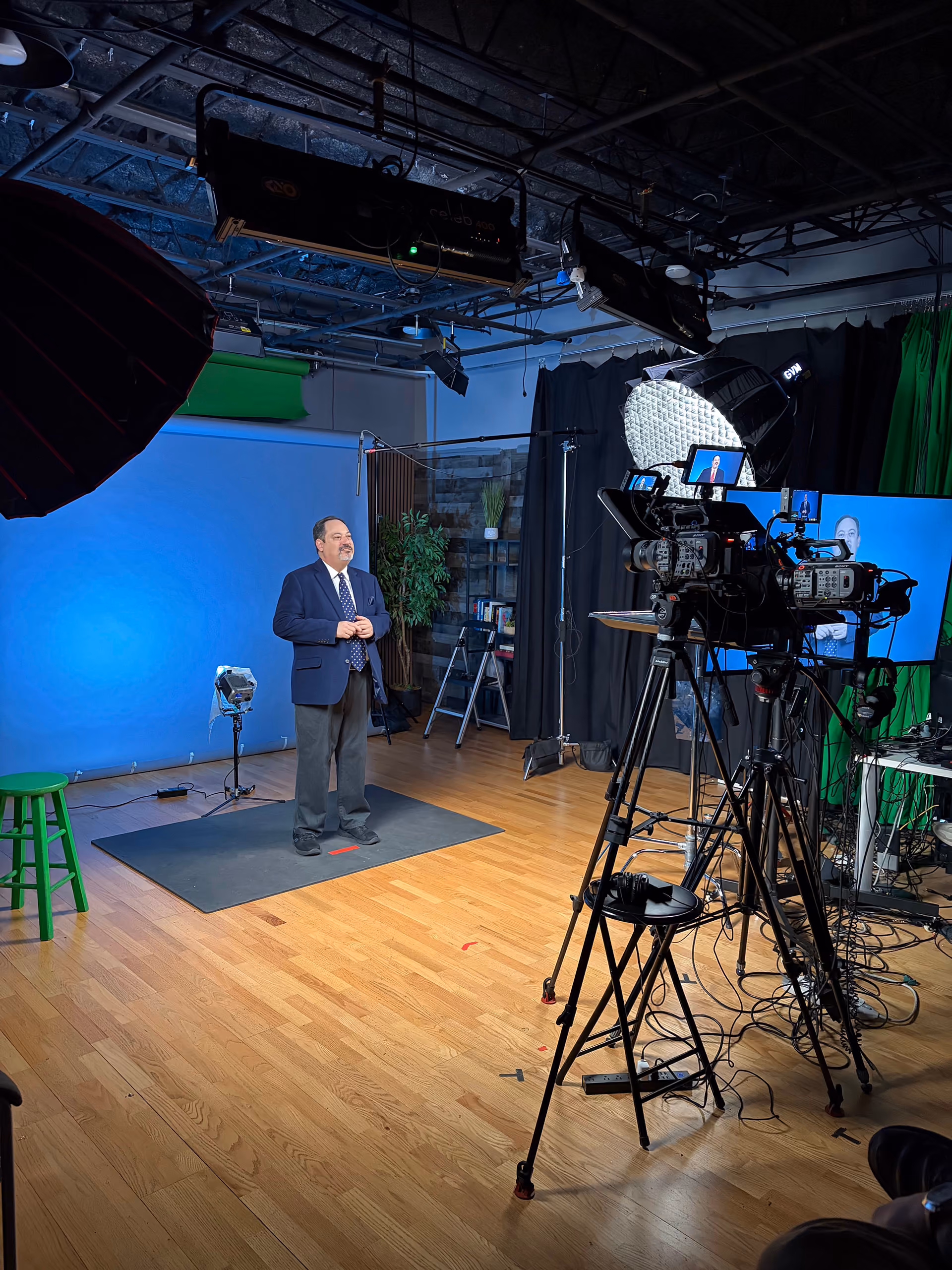 Man in a suit standing on a studio floor marked with red tape in front of a blue backdrop, surrounded by professional video cameras, lighting equipment, and green screen material.