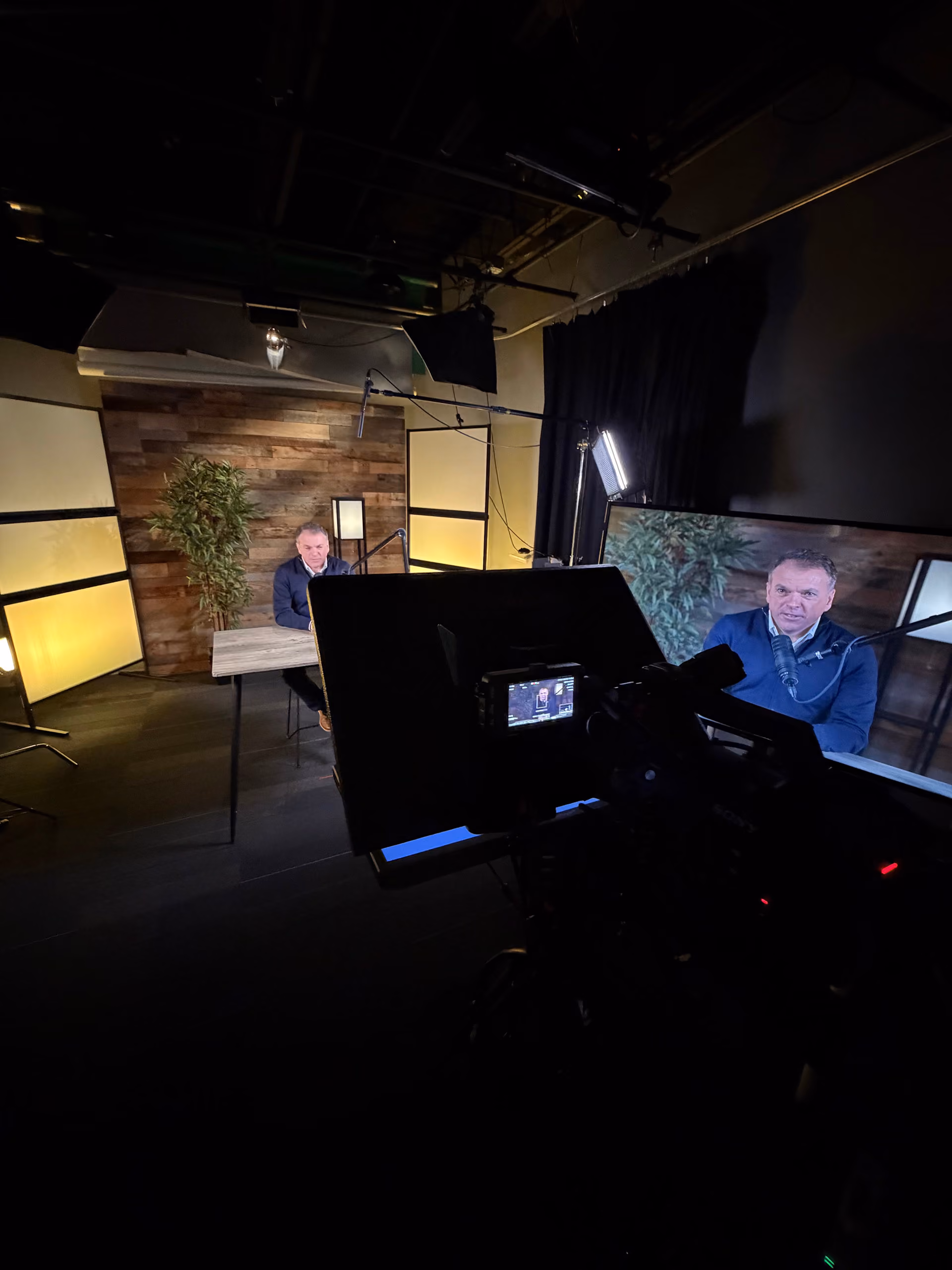 Man sitting at a wooden table in a studio with wood-paneled walls and a potted plant, being filmed by a camera with a large monitor displaying his image.