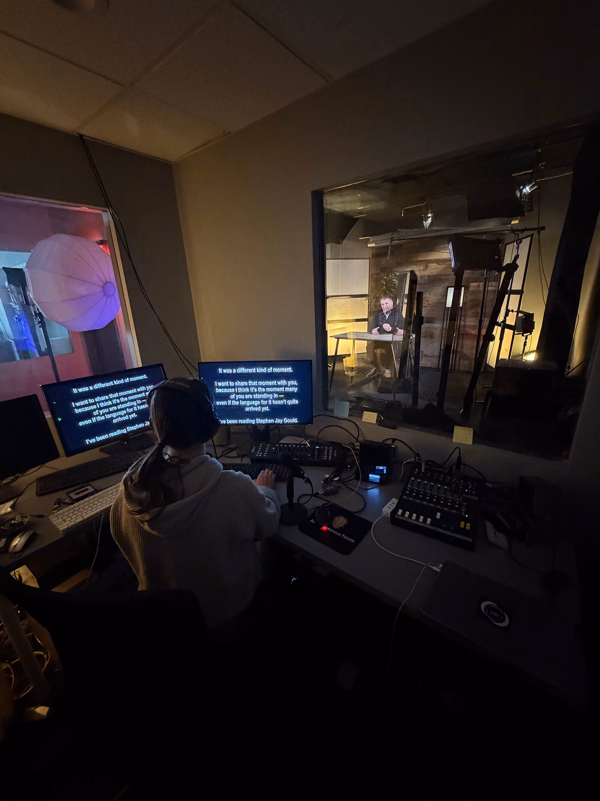 Control room view of a recording studio where a person with headphones operates audio equipment and two screens showing teleprompter text, while another person is visible behind a glass window in a soundproof room with wooden walls and professional lighting.