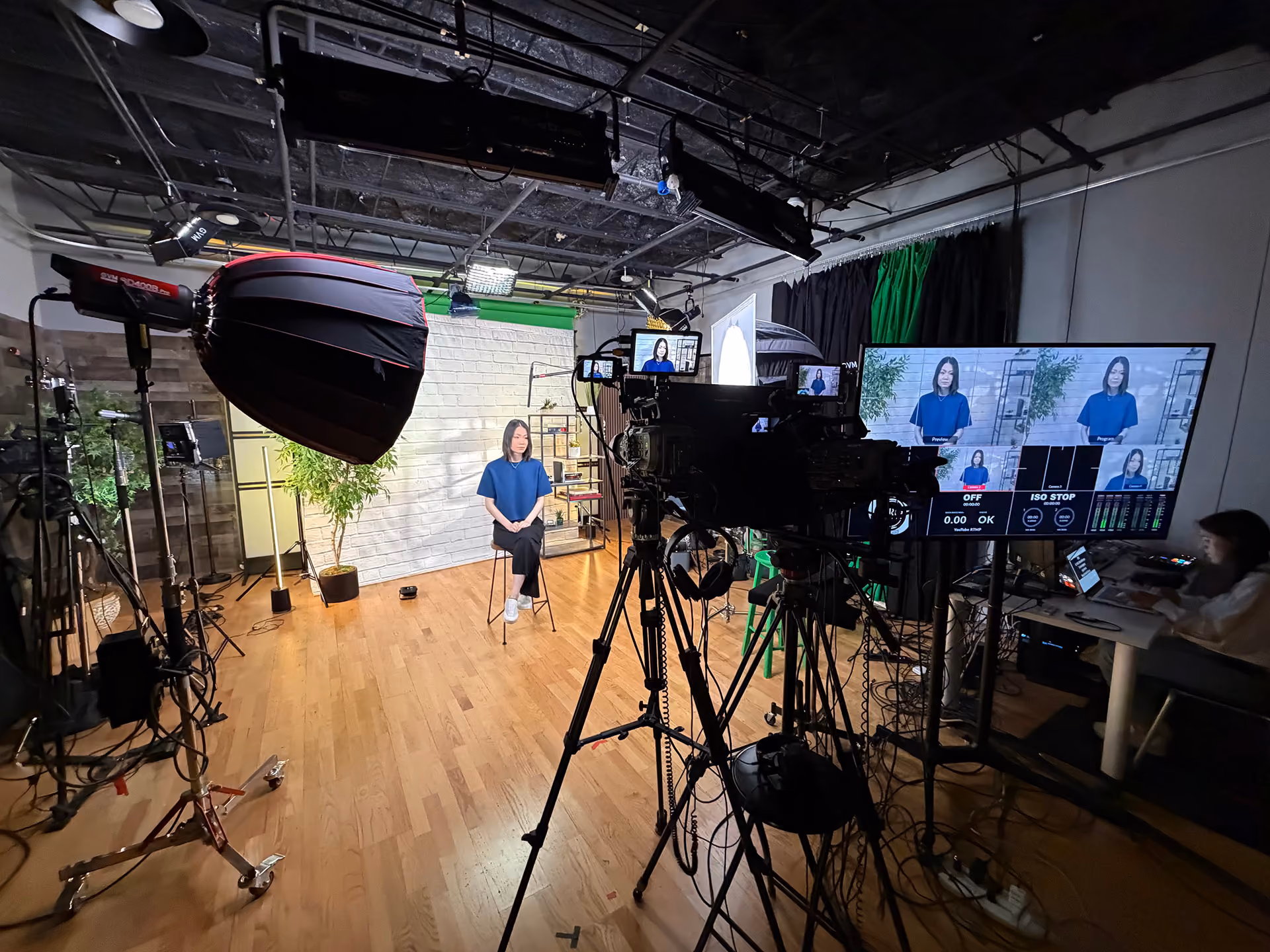 Professional video production studio with a woman seated on a stool in front of a white brick backdrop, surrounded by studio lights, cameras on tripods, and a large monitor showing her live image and camera settings, with a person operating equipment at a desk to the right.