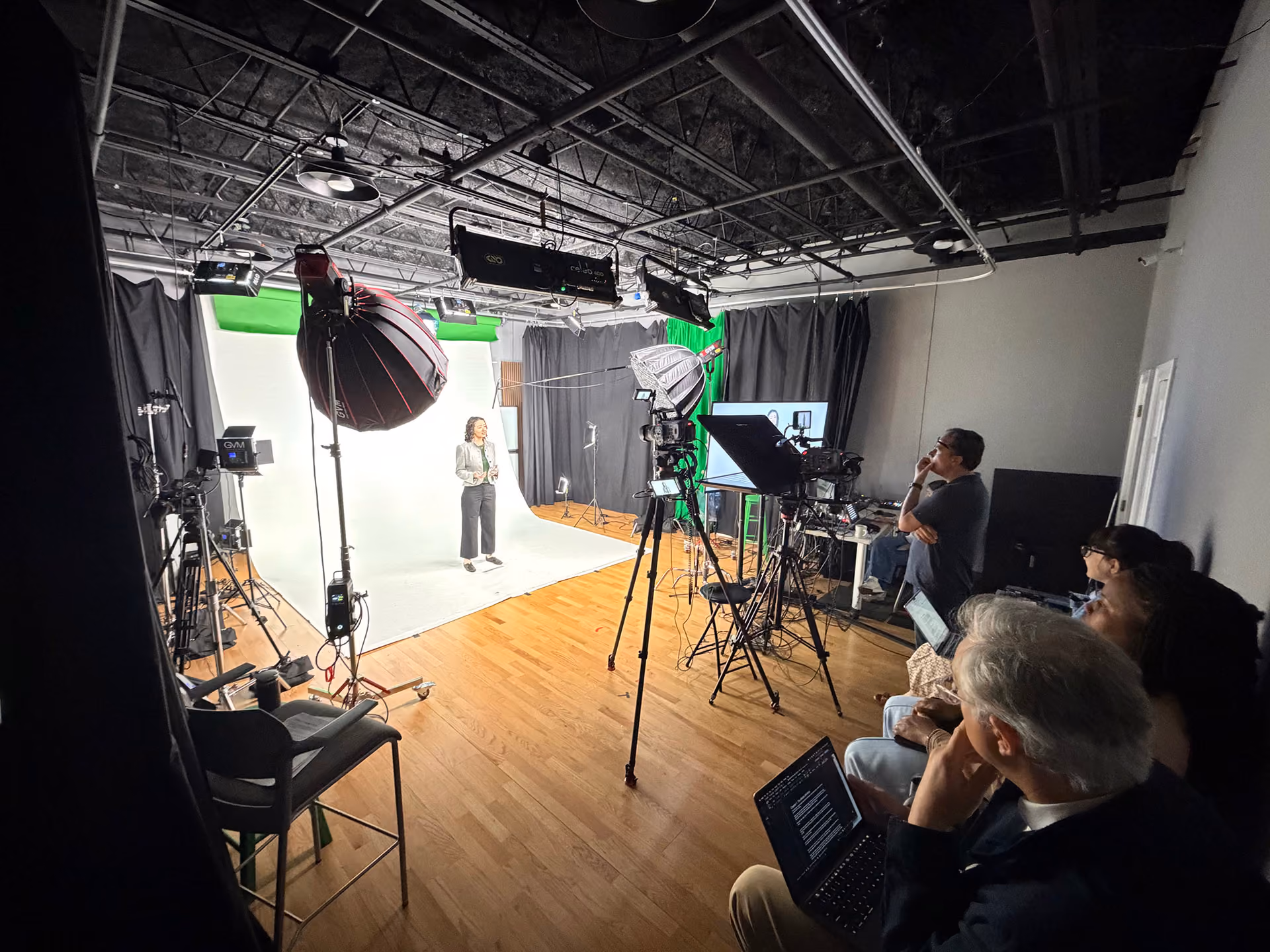 A woman standing in a photography or video studio with a white backdrop and green screen at the top, lit by multiple professional studio lights and surrounded by cameras on tripods, while four people on the right side observe and work on laptops.
